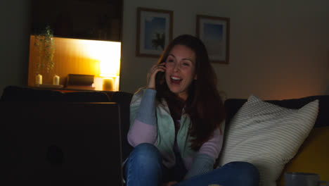 Smiling-Woman-Sitting-On-Sofa-At-Home-At-Night-Talking-On-Mobile-Phone-And-Watching-Movie-Or-Show-On-Laptop-6